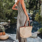 Woman in striped jumpsuit holding woven tote bag by tropical pool, summer fashion outfit