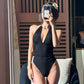 Woman in black halter one-piece swimsuit with high cut legs, taking mirror selfie with flower in hair