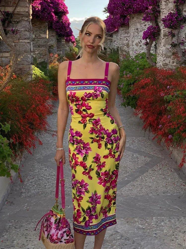 Woman in vibrant yellow floral midi dress with pink straps, holding matching bag, walking through garden path