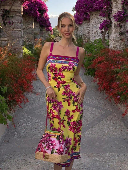 Woman in vibrant yellow floral midi dress with matching bag, standing on stone path surrounded by purple blooms