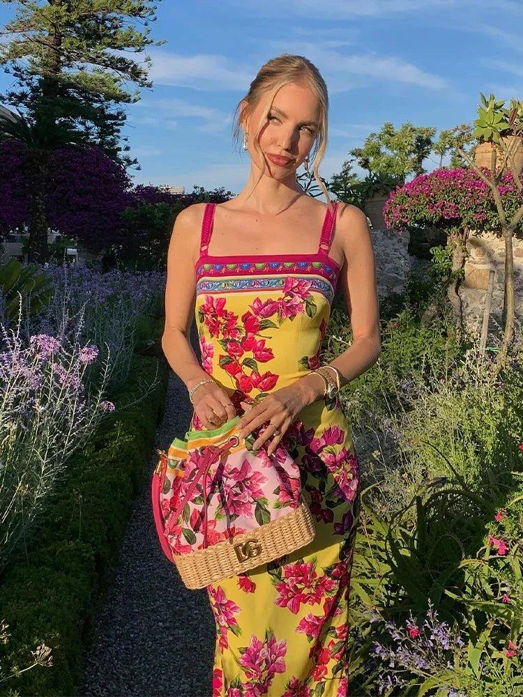 Woman in vibrant floral yellow dress with pink straps, holding woven bag, standing in garden