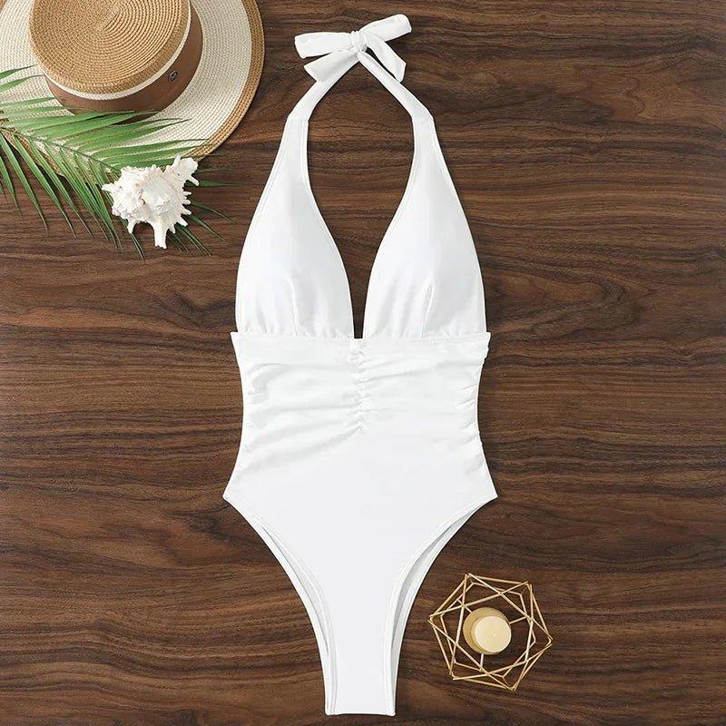 White halter neck one-piece swimsuit with ruched waist, beach hat, palm leaf, and seashell on wood