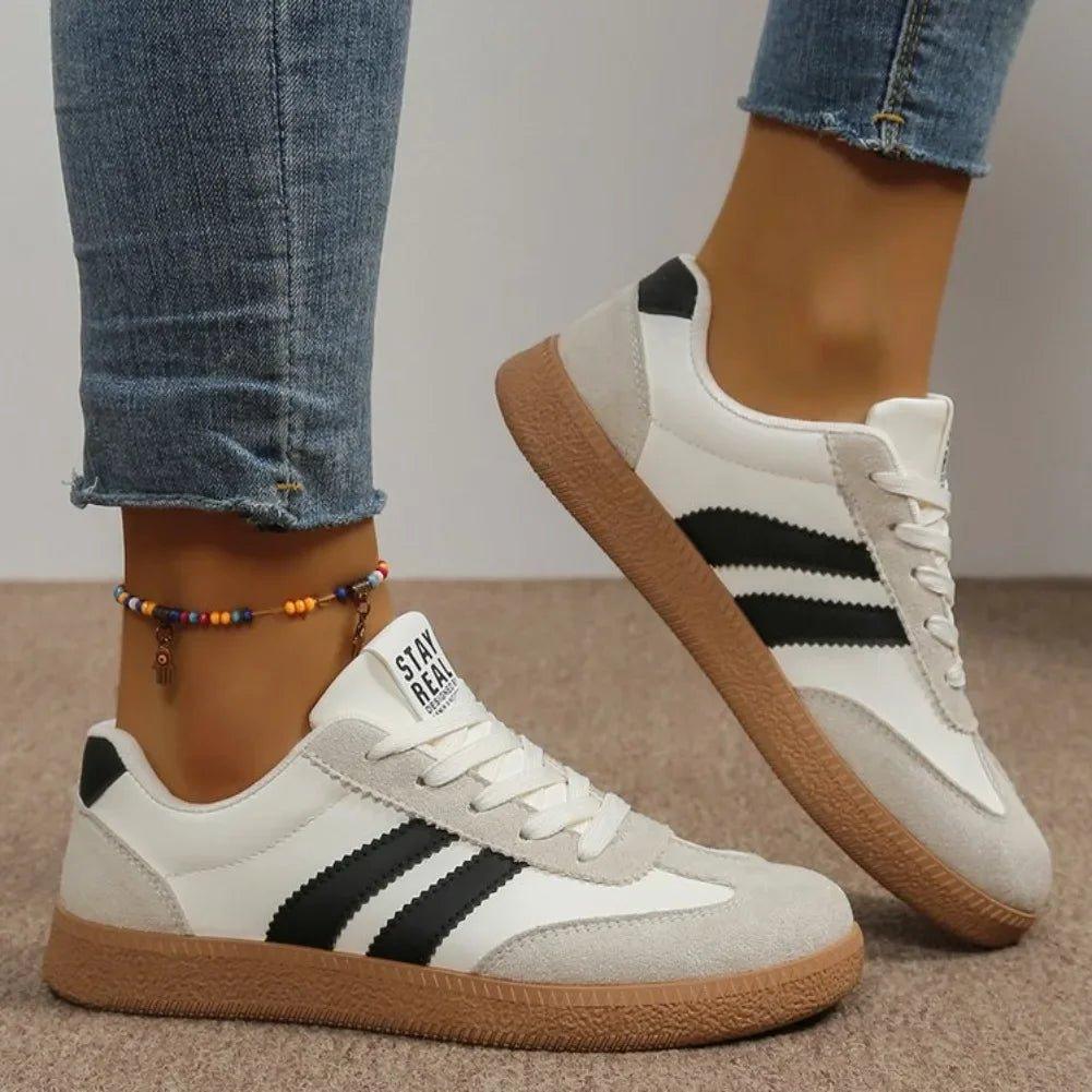 White women's sneakers with black stripes, gum sole, and 'STAY REAL' text, styled with jeans and ankle bracelet