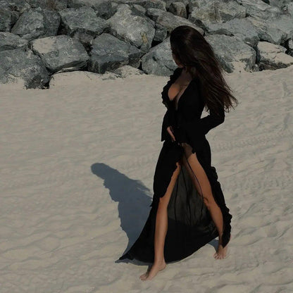 Woman in black beach cover-up dress with high slit walking on sandy shore near rocks