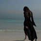 Woman in black flowy dress walking barefoot on beach, ocean view, fashion style
