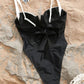 Black one-piece swimsuit with white straps and front bow, styled on sandy beach with butterfly necklace
